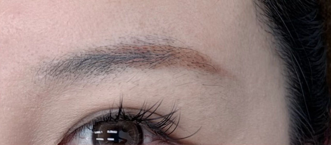 A fourth client before the appointment, photographed close: an older brow tattoo faded to a flat grey with a warm pink edge, sitting outside the natural brow line.
